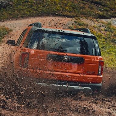 2027 Honda Passport TrailSport Elite in Sunset Orange back splashing through a muddy off-road trail (Mid-Size SUV).
